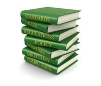 Stack of books labeled "Standards," "Rules," "Regulations," "Policy,""Guidelines," "Directory," and "Compliance"